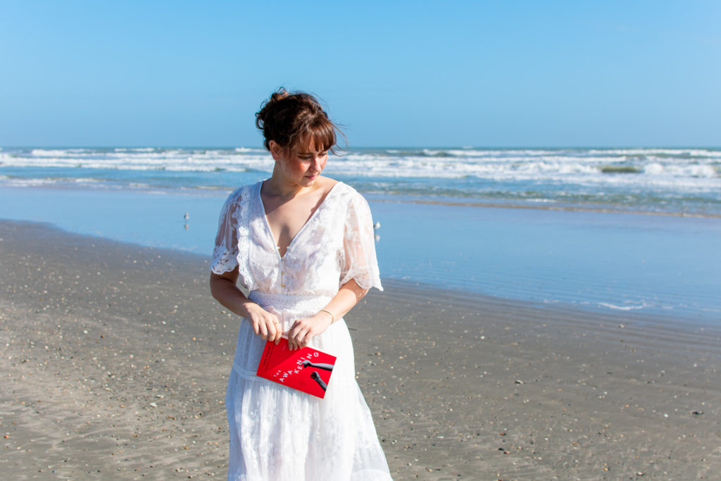 Woman in a white dress standing on the beach with The Awakening by Kate Chopin.