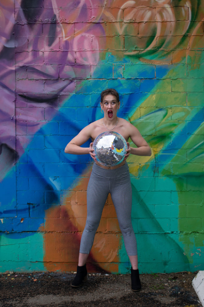 A topless woman in houndstooth pants and black booties, holding a disco ball in front of her.