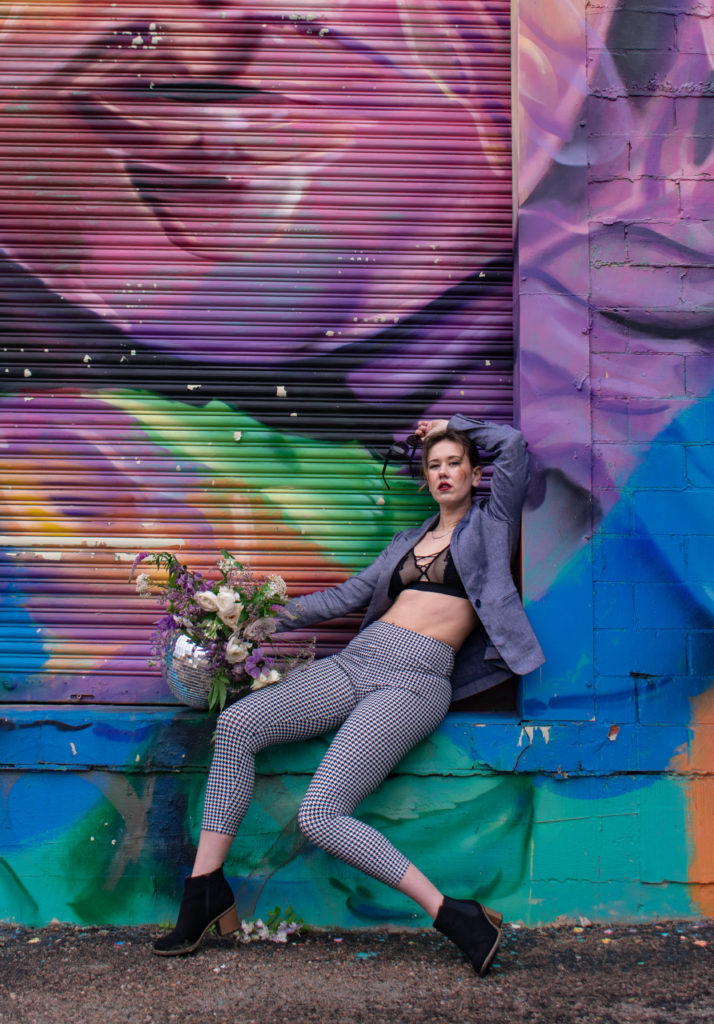 A  woman in houndstooth pants, a black lace bra, blue blazer, and black booties, holding a disco ball covered in flowers in front of a mural.