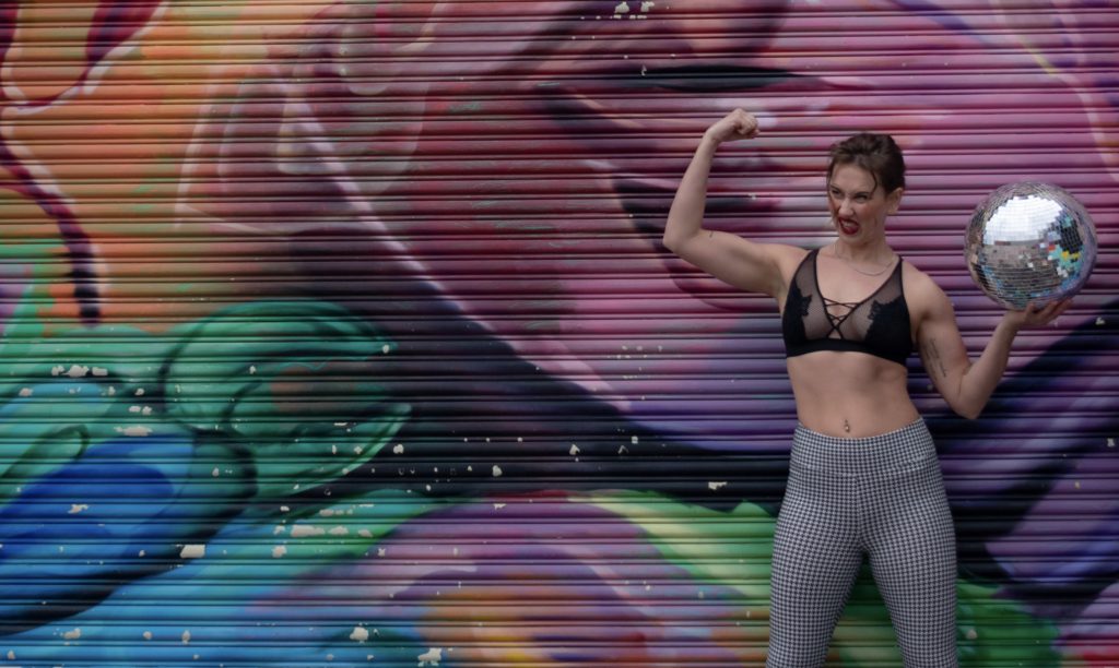 A  woman in houndstooth pants, a black lace bra, and black booties, holding a disco ball, flexing her arm muscles and making a goofy face in front of a mural.