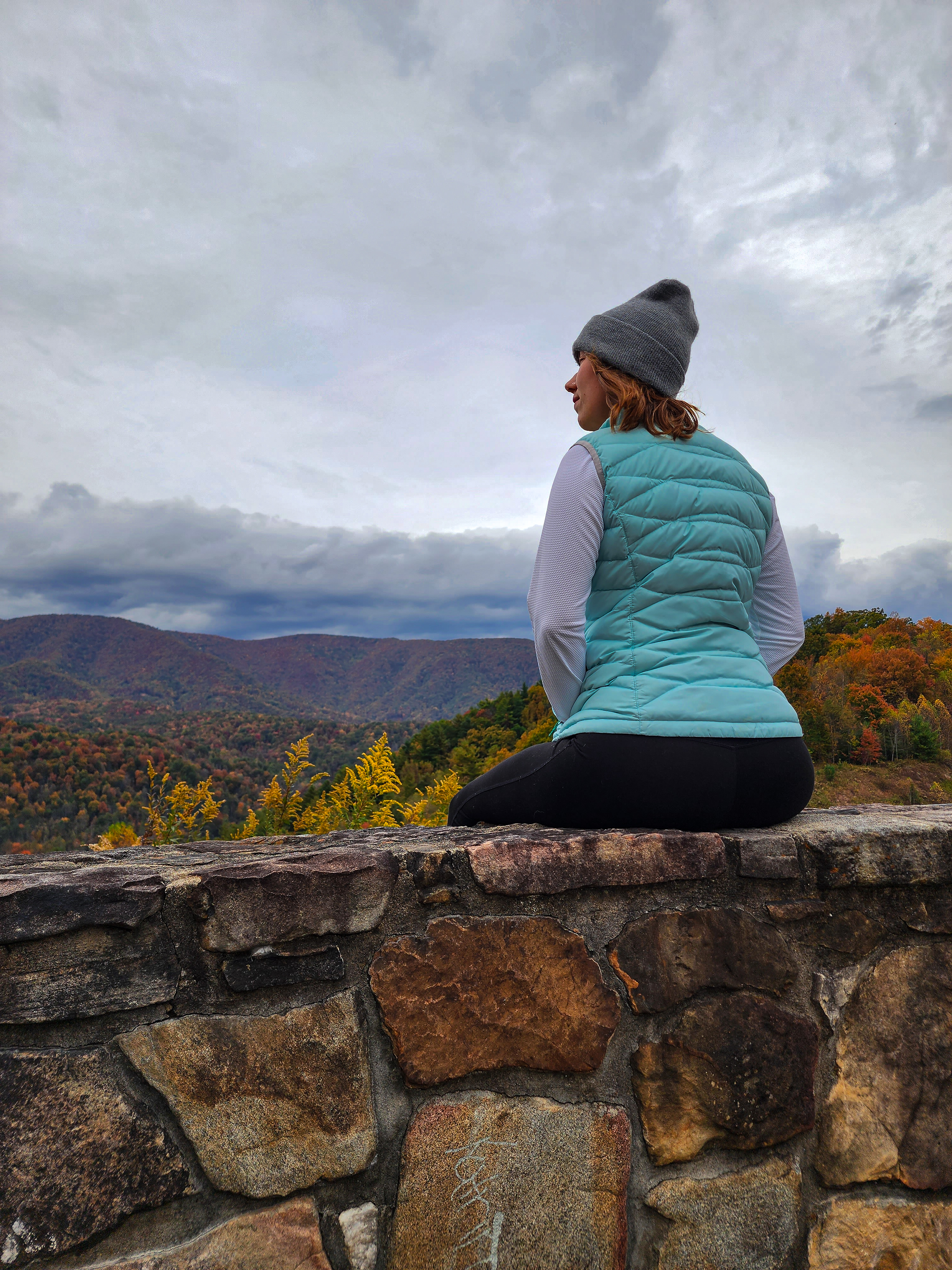 A woman in a vest sitting on the ledge of a scenic overlook in the Appalachia mountains in Tennessee.