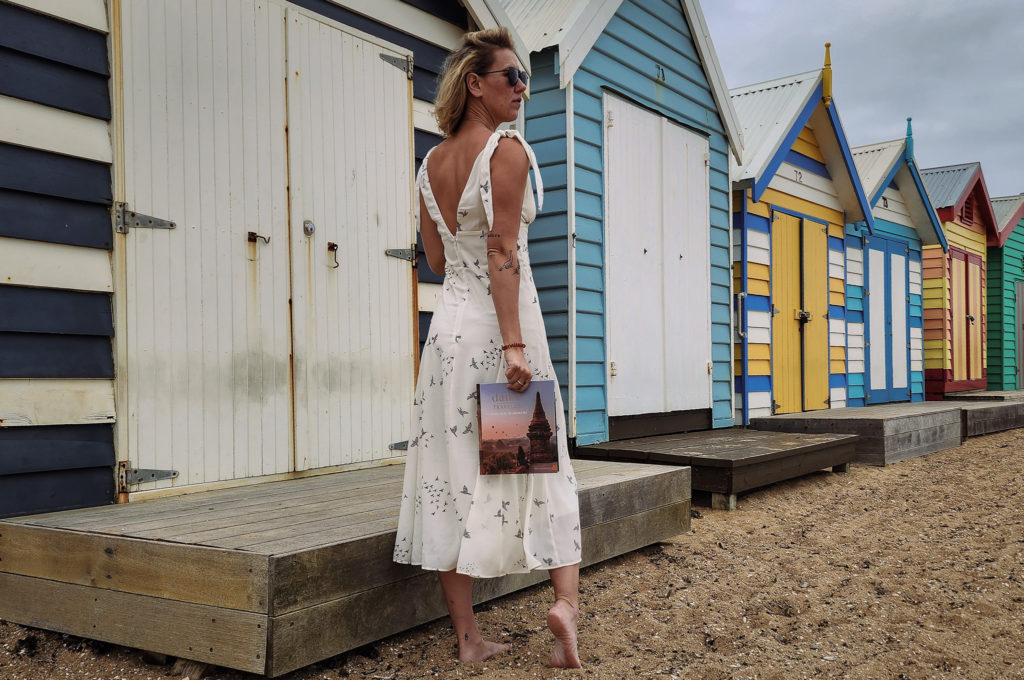 A woman standing on Brighton Beach in Melbourne looking at Bathing Boxes, holding Dame Traveler.