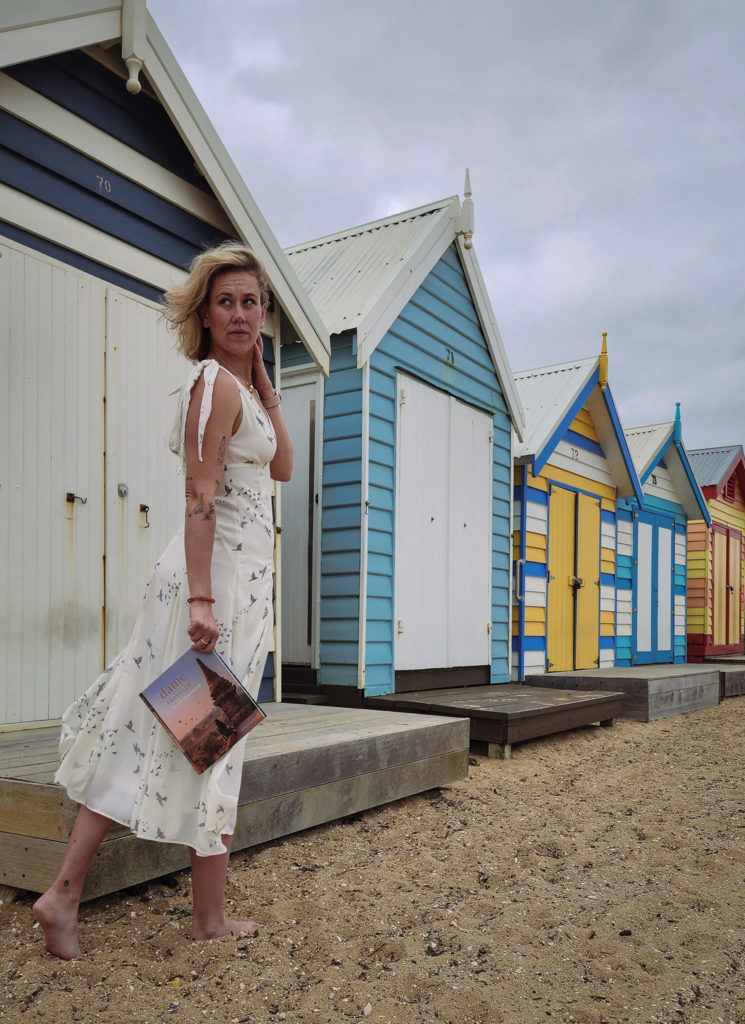 A woman standing on Brighton Beach in Melbourne looking at Bathing Boxes, holding Dame Traveler.